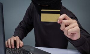 Pessoa mascarada usando computador segura um cartão de crédito dourado, representando uma situação de fraude digital envolvendo cartão de crédito clonado ou roubado.