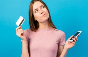 Mulher pensativa segura um celular em uma mão e um cartão de crédito na outra, em frente a um fundo azul, representando dúvidas comuns ao considerar a solicitação de um cartão de crédito para negativados.