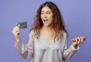 Mulher sorridente segura um smartphone em uma mão e um cartão de crédito na outra, expressando surpresa positiva, em frente a um fundo roxo — representando satisfação ao usar cartões sem anuidade.